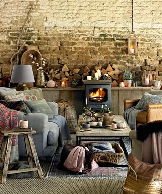 Fall mantel ideas with an exposed stone wall, stove, grey sofas with blankets and firewood and mismatched ornaments on the mantel