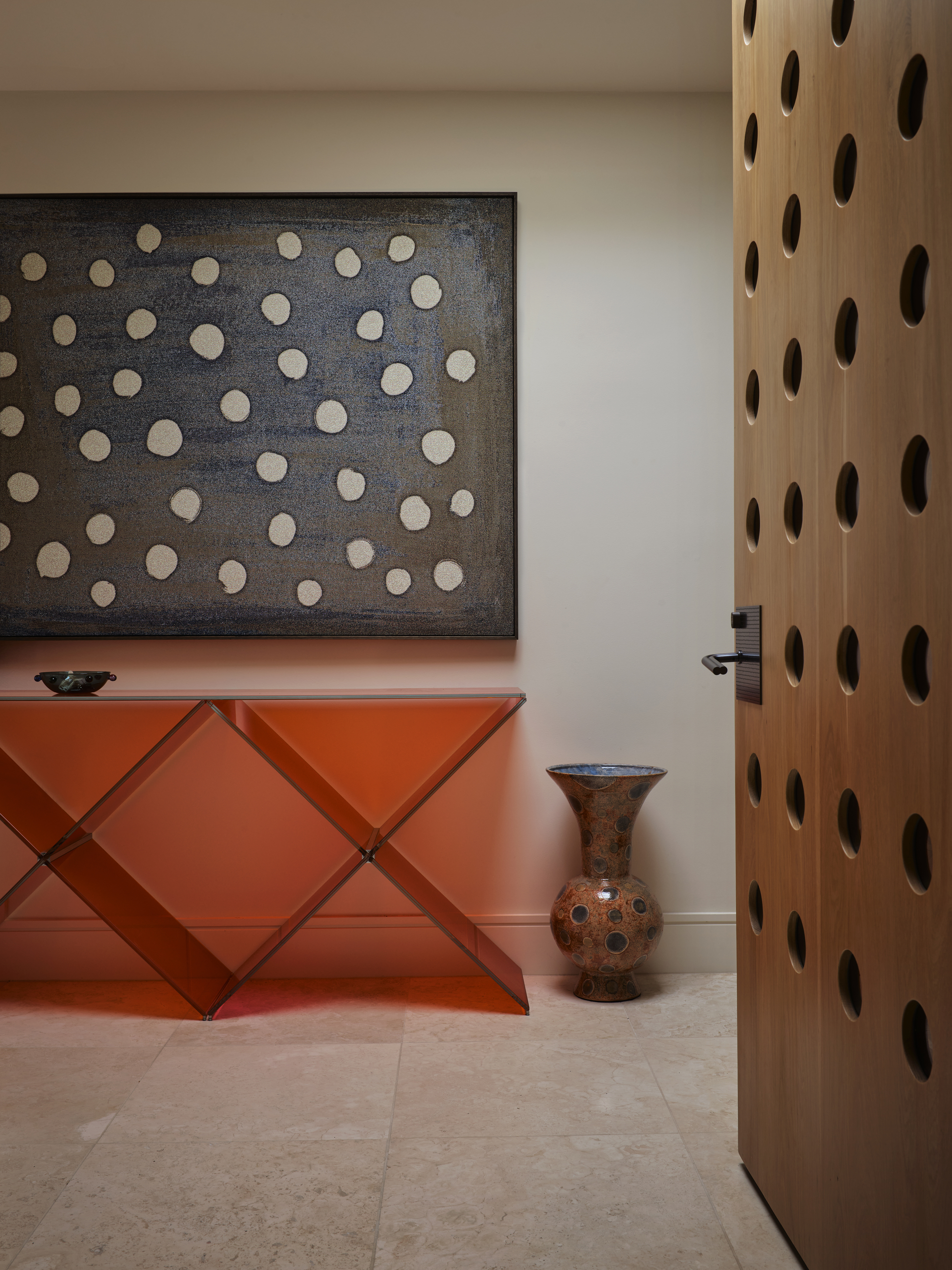 modern entryway with orange perspex console table with polka dot artwork above, a dotted vase on the travertine floor tiles, and a timber door with perforated holes in it