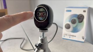 A finger points at the lenses on the small black body of the Piko+ webcam