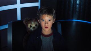 A boy sits in a chair with a teddy bear looking over his shoulder 