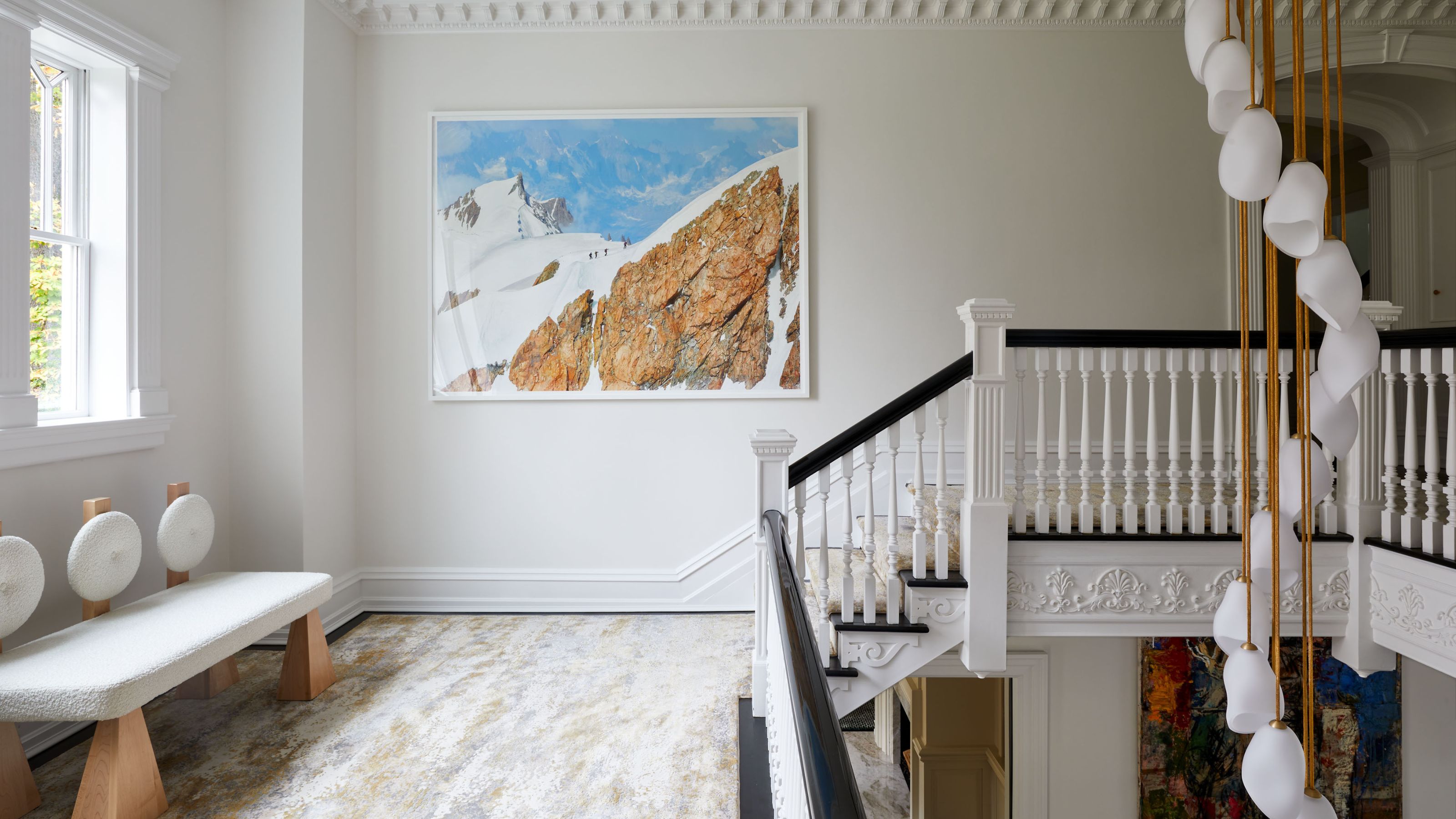 Landing with cream walls and a mountain print on the far wall. A white and wooden bench is on the left hand side and the top of the staircase is on the right. A modern lighting fixture hangs through the opening at the top of the stairs
