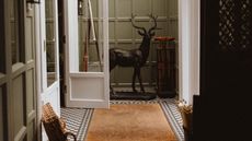 The entrance patio of a country house, with pale green-painted wooden walls, checkered tiled floors in black and white, a rattan rug and a basket, and a deer-shaped sculpture in black bronze placed in the center.
