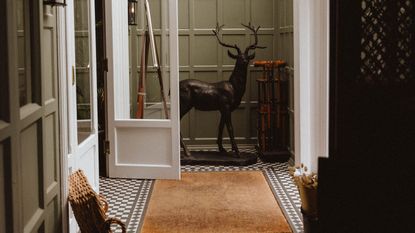 The entrance patio of a country house, with pale green-painted wooden walls, checkered tiled floors in black and white, a rattan rug and a basket, and a deer-shaped sculpture in black bronze placed in the center.