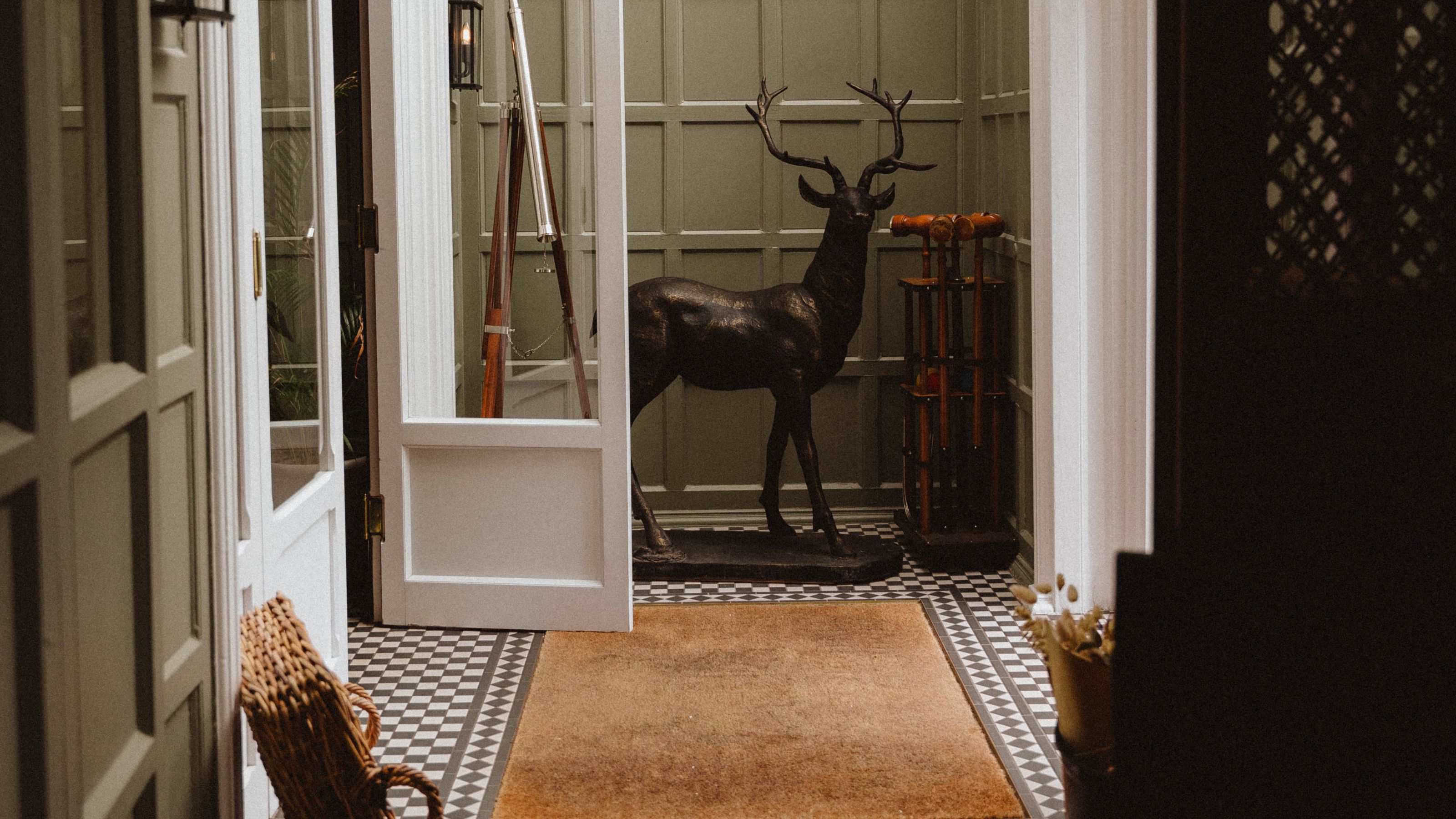 The entrance patio of a country house, with pale green-painted wooden walls, checkered tiled floors in black and white, a rattan rug and a basket, and a deer-shaped sculpture in black bronze placed in the center.
