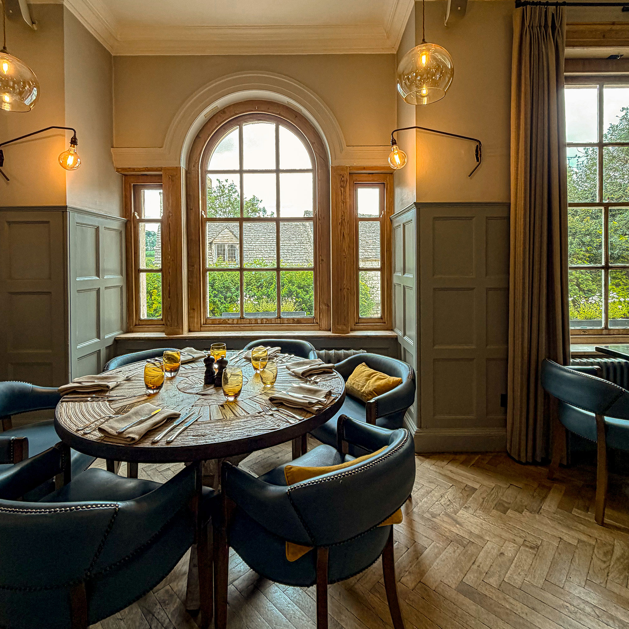 Dining room with wooden herringbone flooring, half panelled walls and round dining table surrounded by leather upholstered chairs