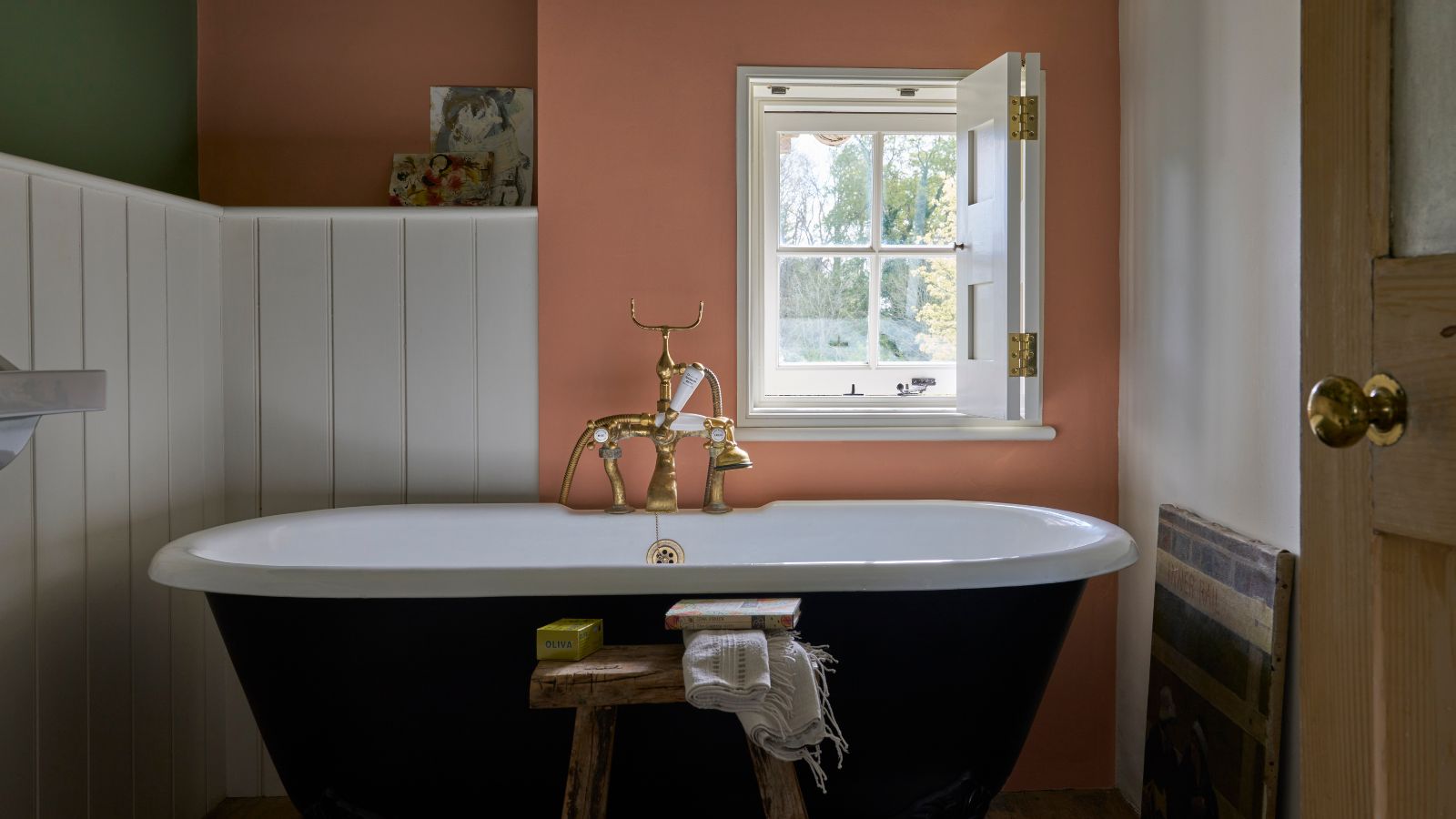 A cottage bathroom with terracotta walls, a roll-top bath, white panelled wall, and a small window. 