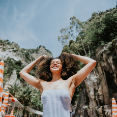 A young, mixed race woman in a striped bathing costume stands on a beautiful Italian beach on a sunny day. She raises her arms and runs her fingers through her hair. She looks serene, confident, satisfied, and pleased to be enjoying a European vacation.
