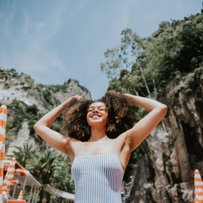 A young, mixed race woman in a striped bathing costume stands on a beautiful Italian beach on a sunny day. She raises her arms and runs her fingers through her hair. She looks serene, confident, satisfied, and pleased to be enjoying a European vacation.