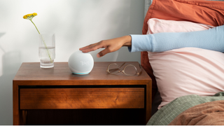 En Echo Dot-smarthögtalare med klocka står på ett bord bredvid en säng.