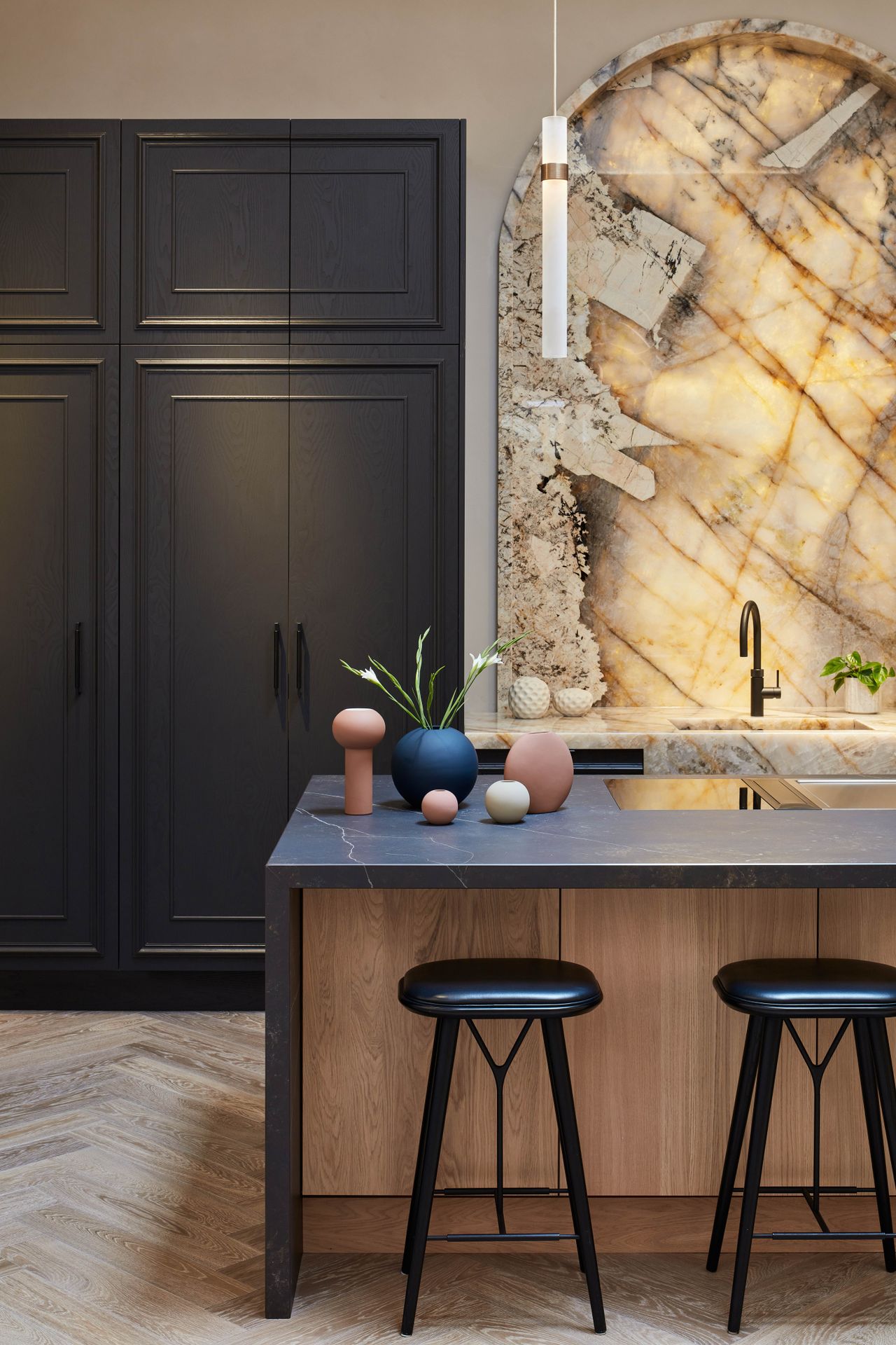 a large, arched backlit onyx stone backsplash in a kitchen above the sink and in front of the kitchen island with black stool seating and tall black cabinetry