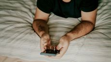 A man lying on a bed holds a smartphone, only his hands and upper torso showing. 
