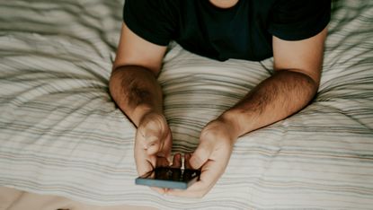 A man lying on a bed holds a smartphone, only his hands and upper torso showing. 