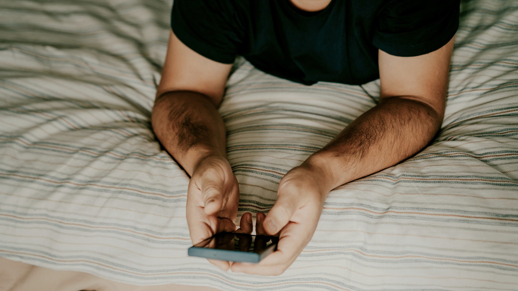A man lying on a bed holds a smartphone, only his hands and upper torso showing. 