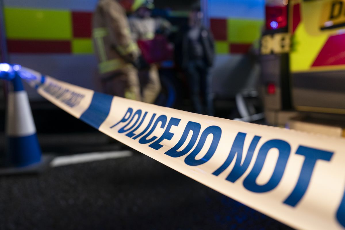 A still from Casualty of police tape reading &quot;POLCE DO NOT&quot; in blue print on a white background fastened to a blue and white traffic cone with ambulances and emergency service people in the background. 