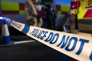 A still from Casualty of police tape reading "POLCE DO NOT" in blue print on a white background fastened to a blue and white traffic cone with ambulances and emergency service people in the background.