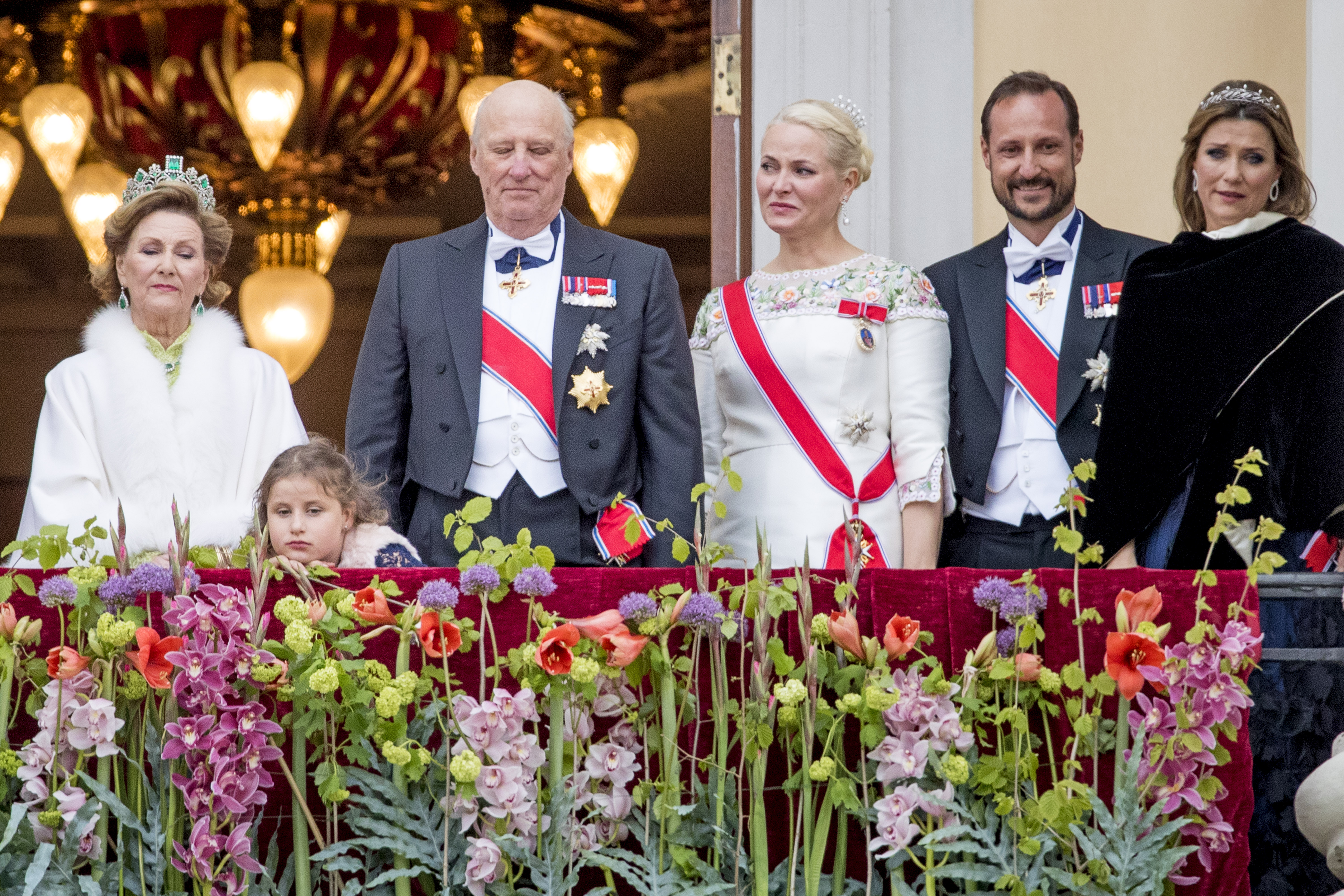 King Harald, Queen Sonia, Crown Prince Haakon, Crown Princess Mette-Marit, Princess Martha Louise smiling on a balcony covered in flowers