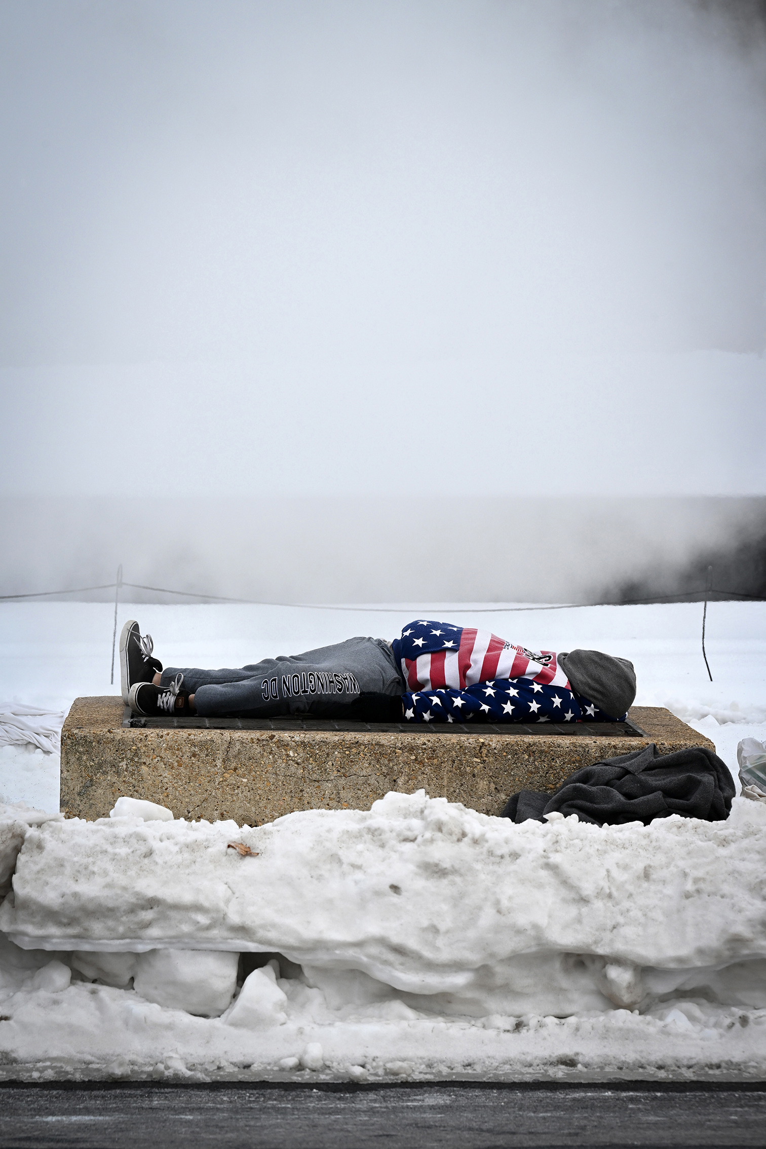 Person bundled in an American flag blanket lies on a concrete platform amid snow and fog