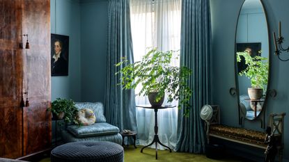 Bedroom with pale blue walls, green carpet, mahogany wardrobe, blue daubed and vintage paintings and wrought iron mirror and table