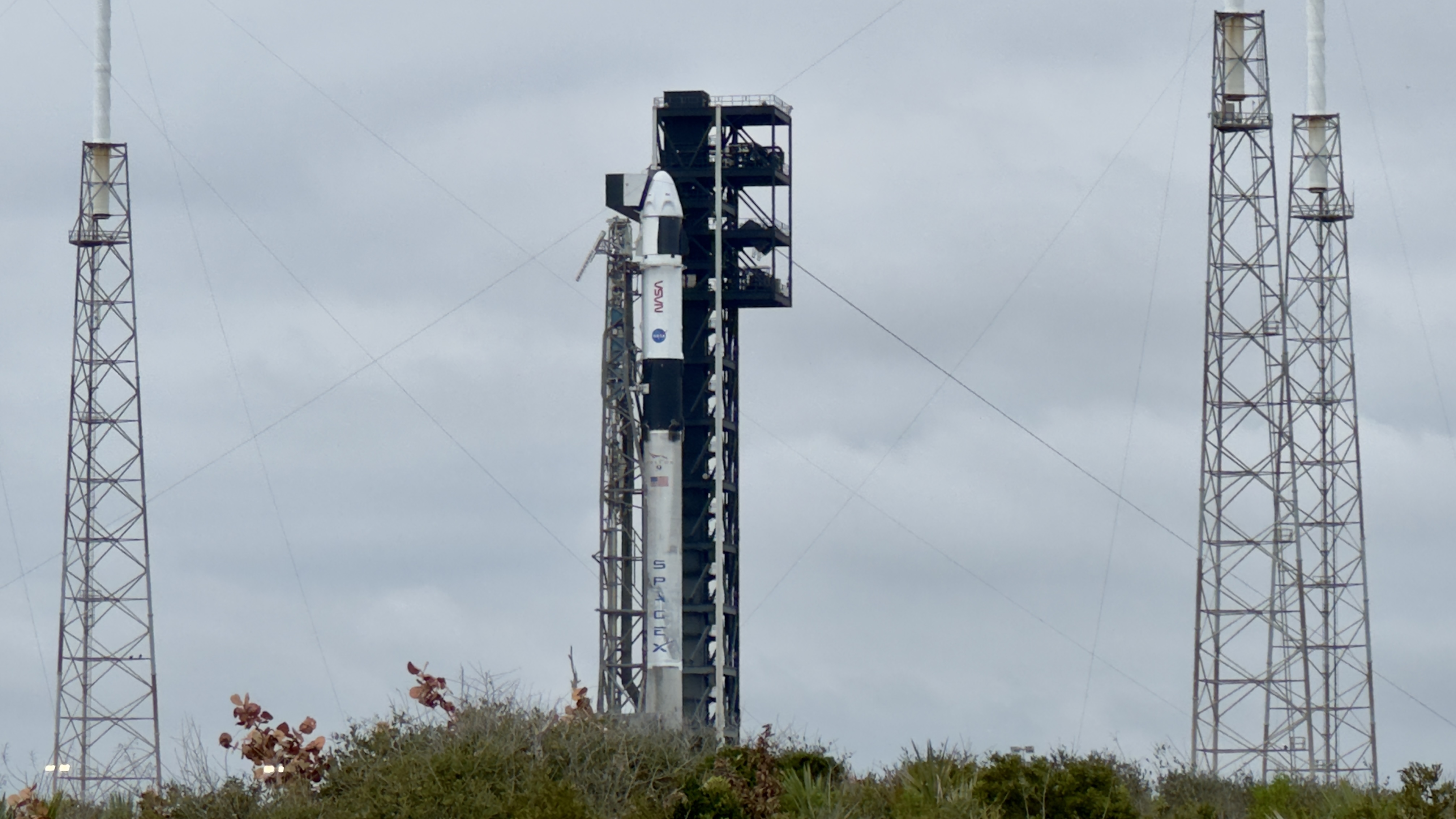 a white rocket stands at a black tower against a grey sky.