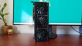 AMD Radeon RX 9070 Asus Prime graphics card standing upright in front of gaming monitor on woodgrain desk.