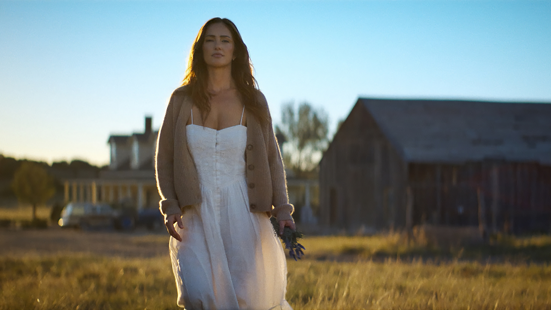 Minka Kelly as Quinn, wearing a white dress and cardigan, holds flowers as she walks through a field with a farmhouse behind her, in Episode 202 of Netflix's 'Ransom Canyon.'