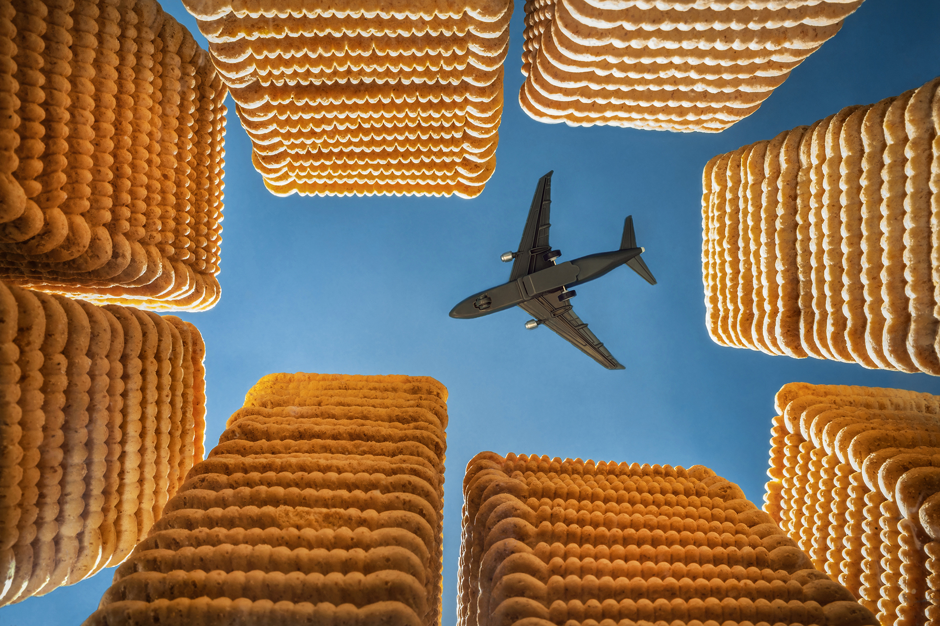 An airplane flies overhead in a blue sky, framed by tall stacks of crackers resembling skyscrapers