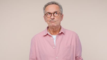 A conflicted-looking older man in a pink shirt against a dusky pink background.