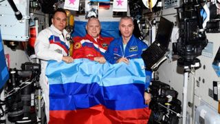 three cosmonauts holding a red, dark-blue, and light-blue-striped flag aboard the international space station
