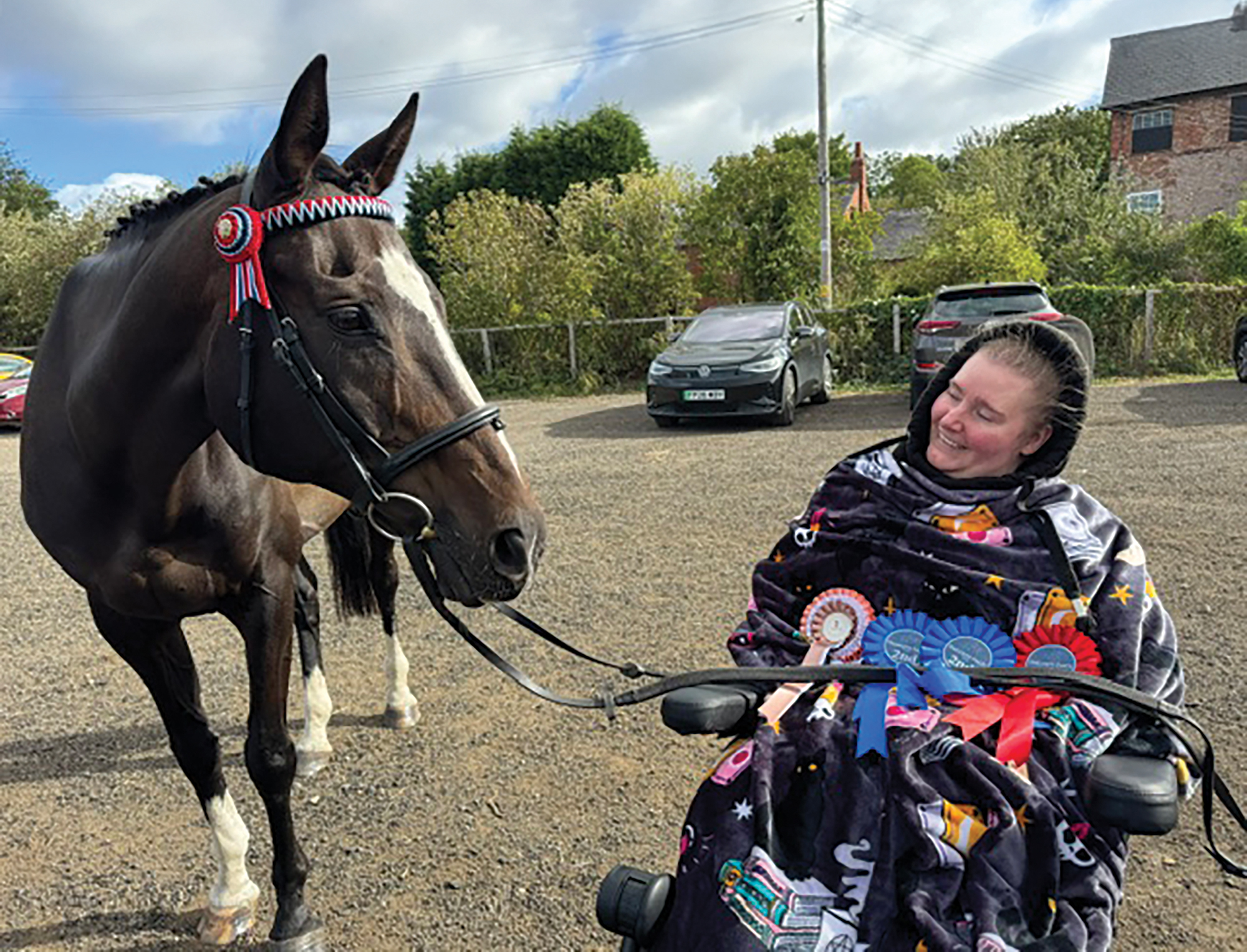 A woman in a wheelchair looks adoringly at a bay horse wearing a bridle with a red, white and blue browband. The horse's reins lay across the lady's lap, where she also has a number of rosettes