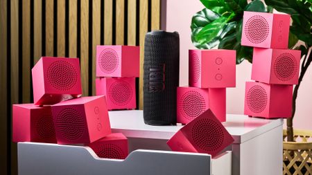 The JBL Flip 7 standing on its end surrounded by piles of pink IKEA Kallsup speakers.