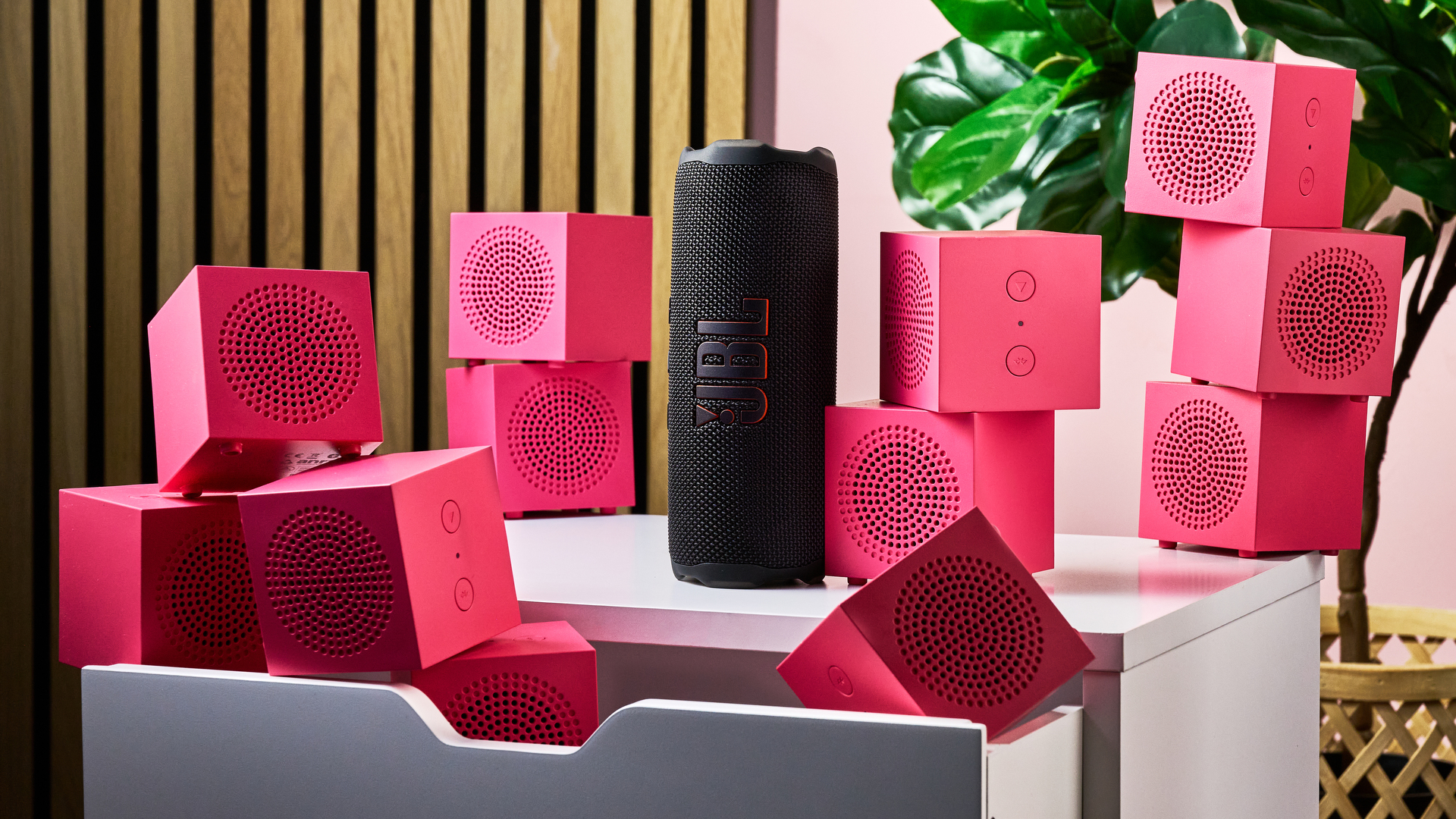 The JBL Flip 7 standing on its end surrounded by piles of pink IKEA Kallsup speakers.