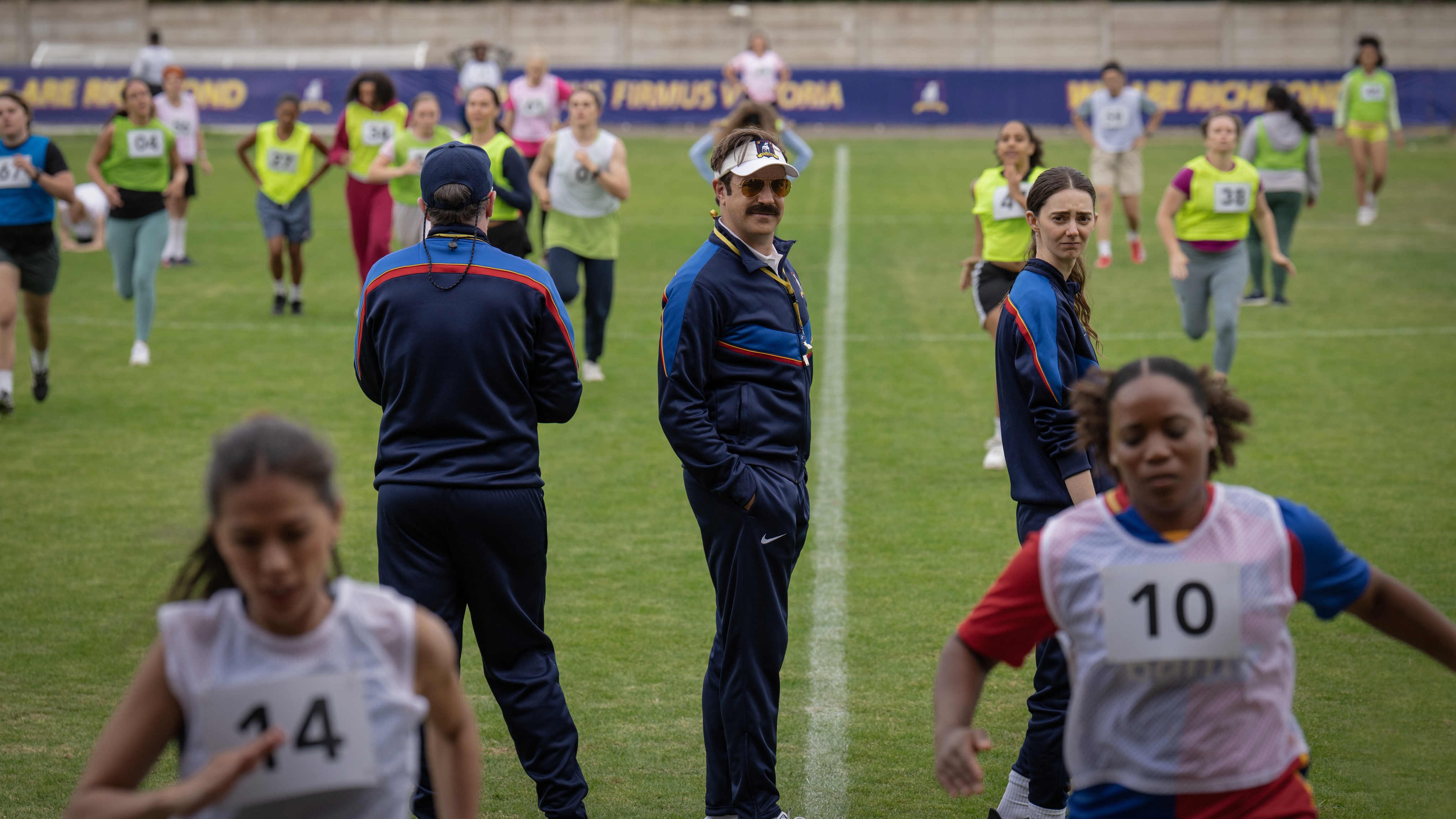 Brendan Hunt as Coach Beard, Jason Sudeikis as Ted Lasso and Tanya Reynolds as Coach Chilton standing on a football pitch coaching a women's football team.