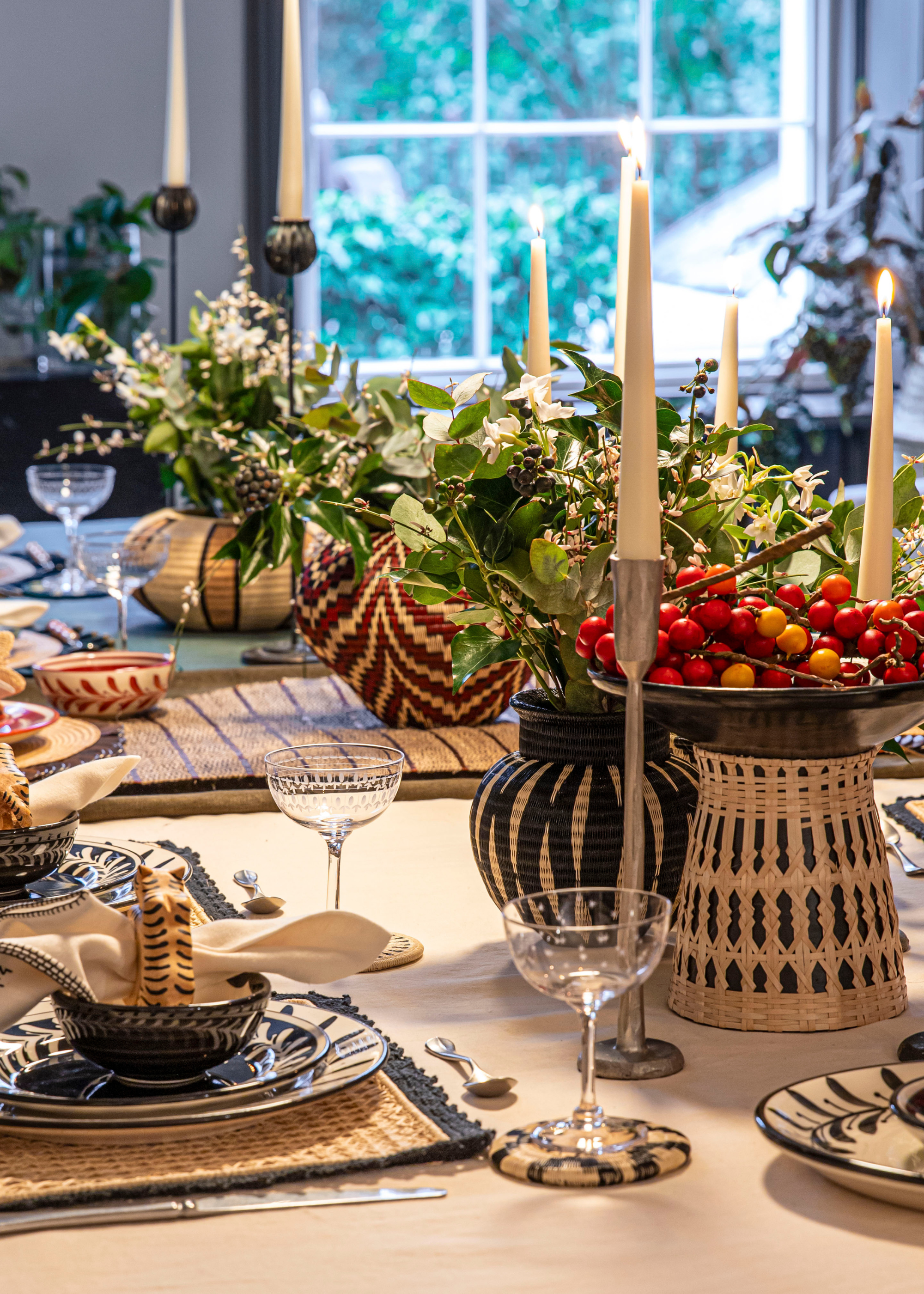 A dining table set with pedestal bowls of fruit, vases of cut stems, tapered candles, and piled plates and napkins