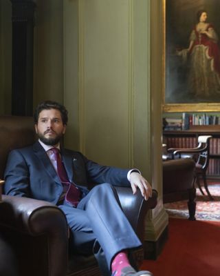 Henry (Kit Harington) wearing a suit, sits in an expensive looking leather armchair in an opulent room, with a portrait of an elegant looking woman from many years ago on the wall behind him