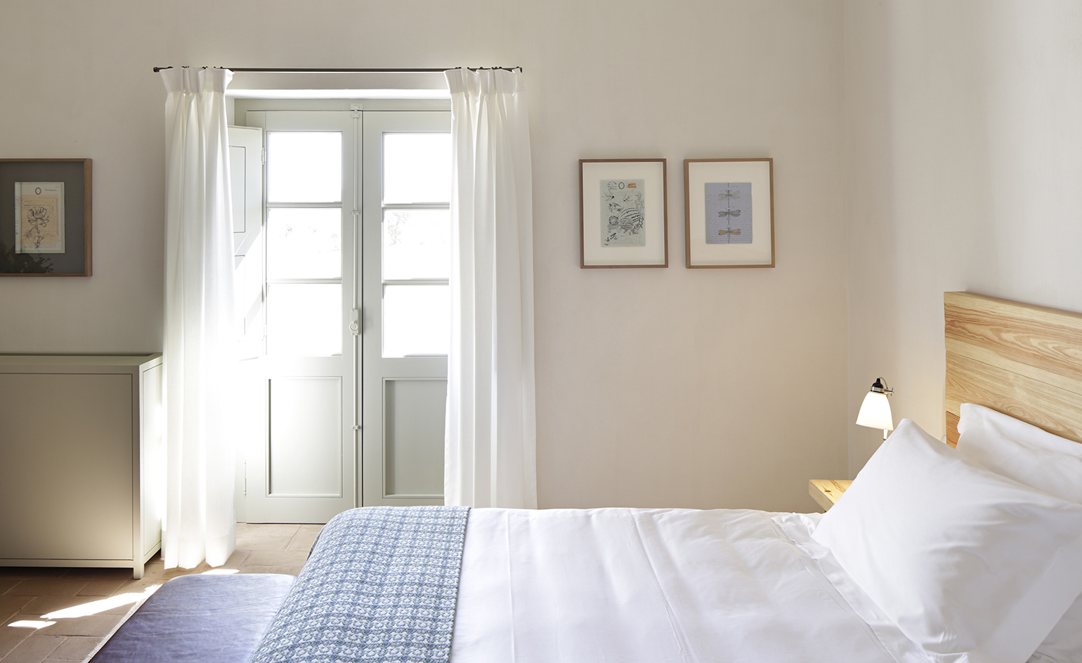 A light and airy room in the São Lourenço do Barrocal hotel. White walls with sketch art of nature on the walls. Wooden bed frame and white linen. To the fartherst wall, there is a balcony door which lets through a lot of light.