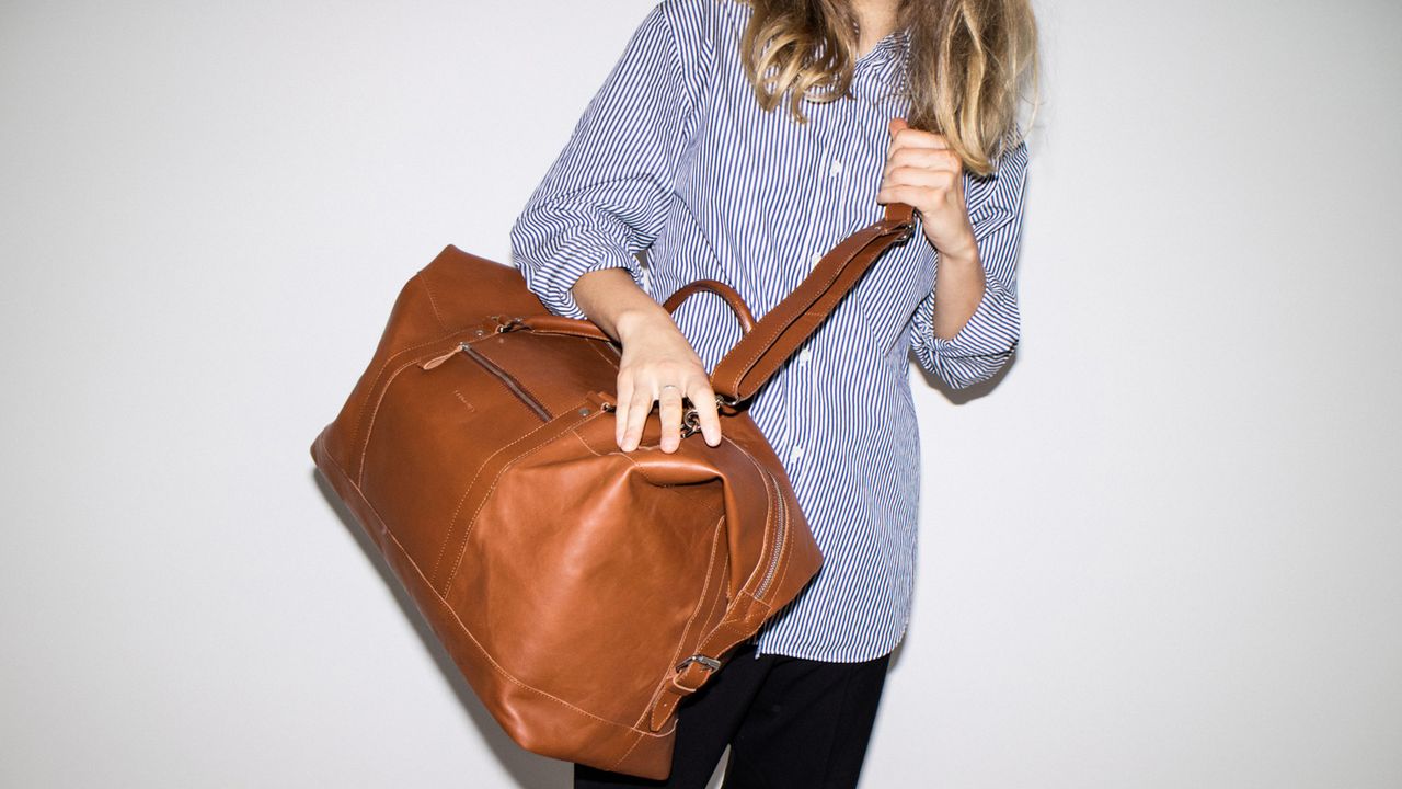 A woman holding one of the best travel bags standing against a white background. It&#039;s tan leather.