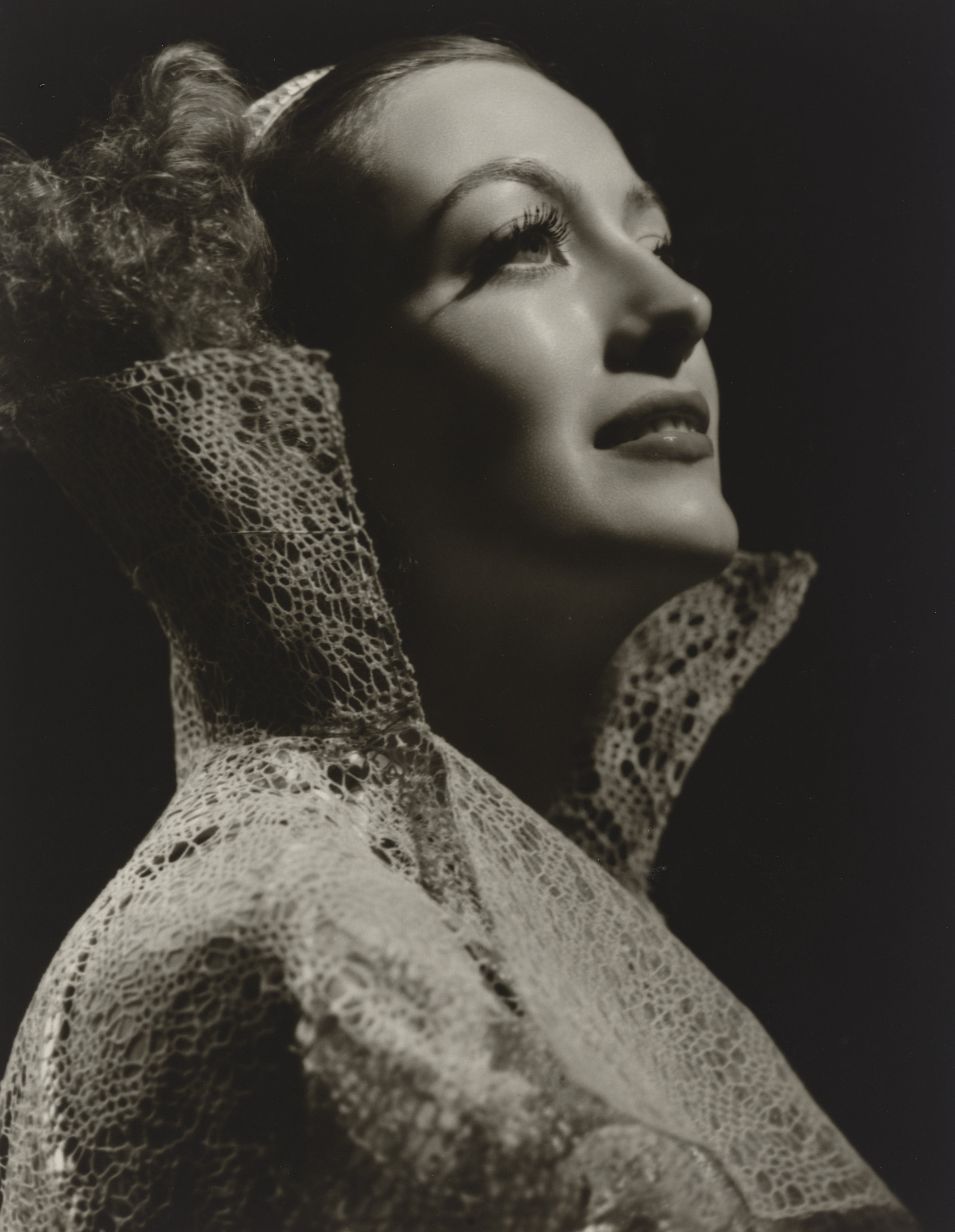 Joan Crawford in profile gazes upwards with a slight smile, her face lit from above against a black background and framed by a tall, stiff collar of open lace.