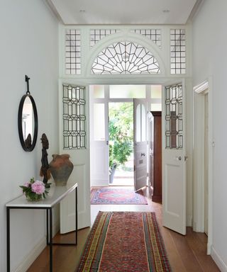 A bright entryway with an open front door filled with natural light with a glass pane arch and doors, two patterned rugs, a white side table and a circular black mirror with pink flowers, a rustic vase and abstract statue