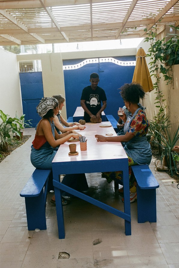 An outdoor patio decorated with blue-painted wooden furniture and a gate in the same hue hosts a ceramic workshop with women working the clay at a table in denim aprons.