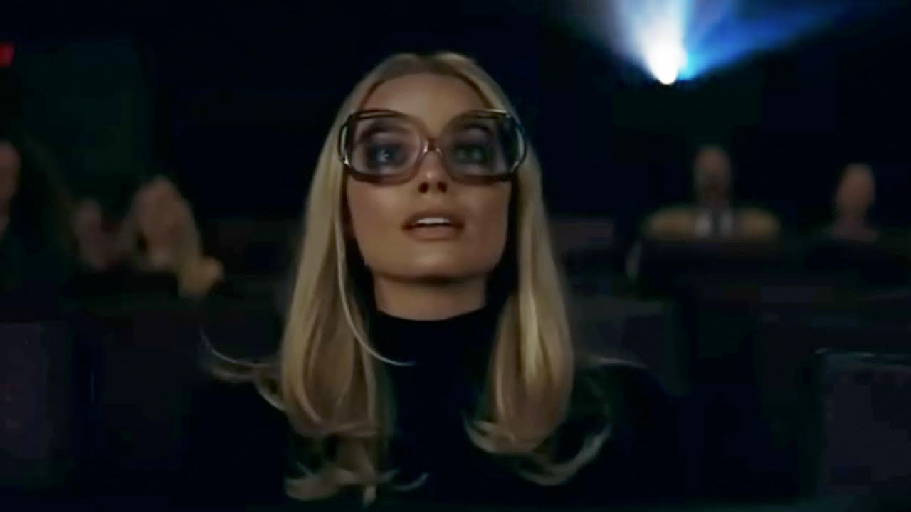 Margot Robbie with huge glasses, sitting by herself in a darkened movie theater. 