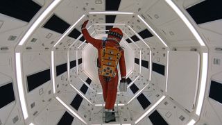 Still from the movie " 2001: A Space Odyssey." An astronaut wearing a dark orange spacesuit is walking through a white and black octagonal-shaped corridor with bright white lights.