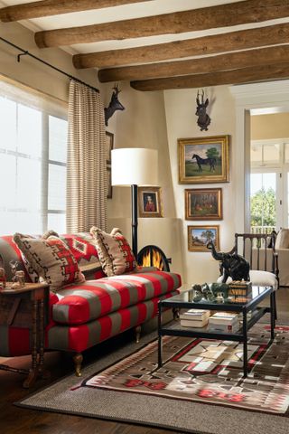Living room with red and gray stiped sofa, patterned rug, glass and metal coffee table, wooden beams, artwork on cream walls