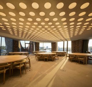 Fjordenhus, by Olafur Eliasson and Studio Olafur Eliasson in Vejle, Denmark. A dining room with round tables, chairs, round roof lights and large windows overlooking a body of water.