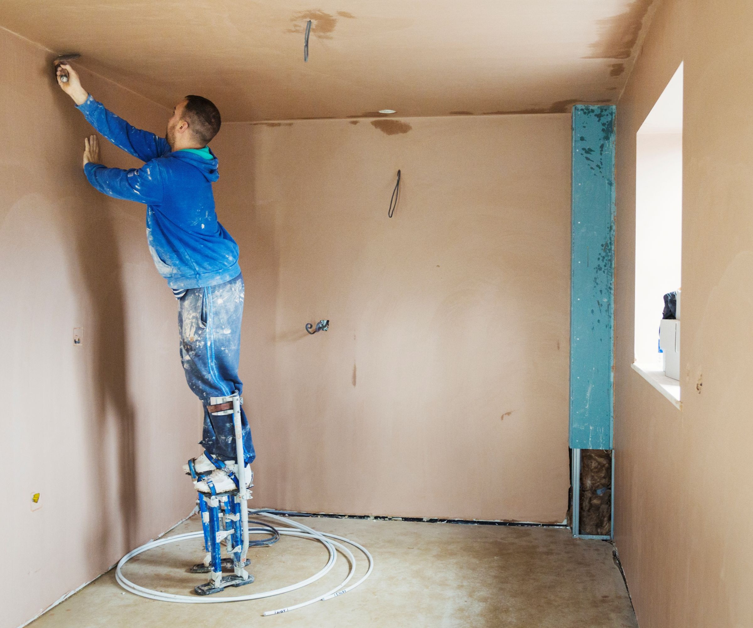 A man in blue clothing on stilts plastering a wall