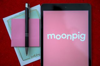 A decorative image for TechRadar's Moonpig discount codes page. A birthday card with a pink envelope sat on top of it. On top of the envelope is a black pen and to the side of this stationery is an iPad with the Moonpig logo displayed on it on a pink background. 