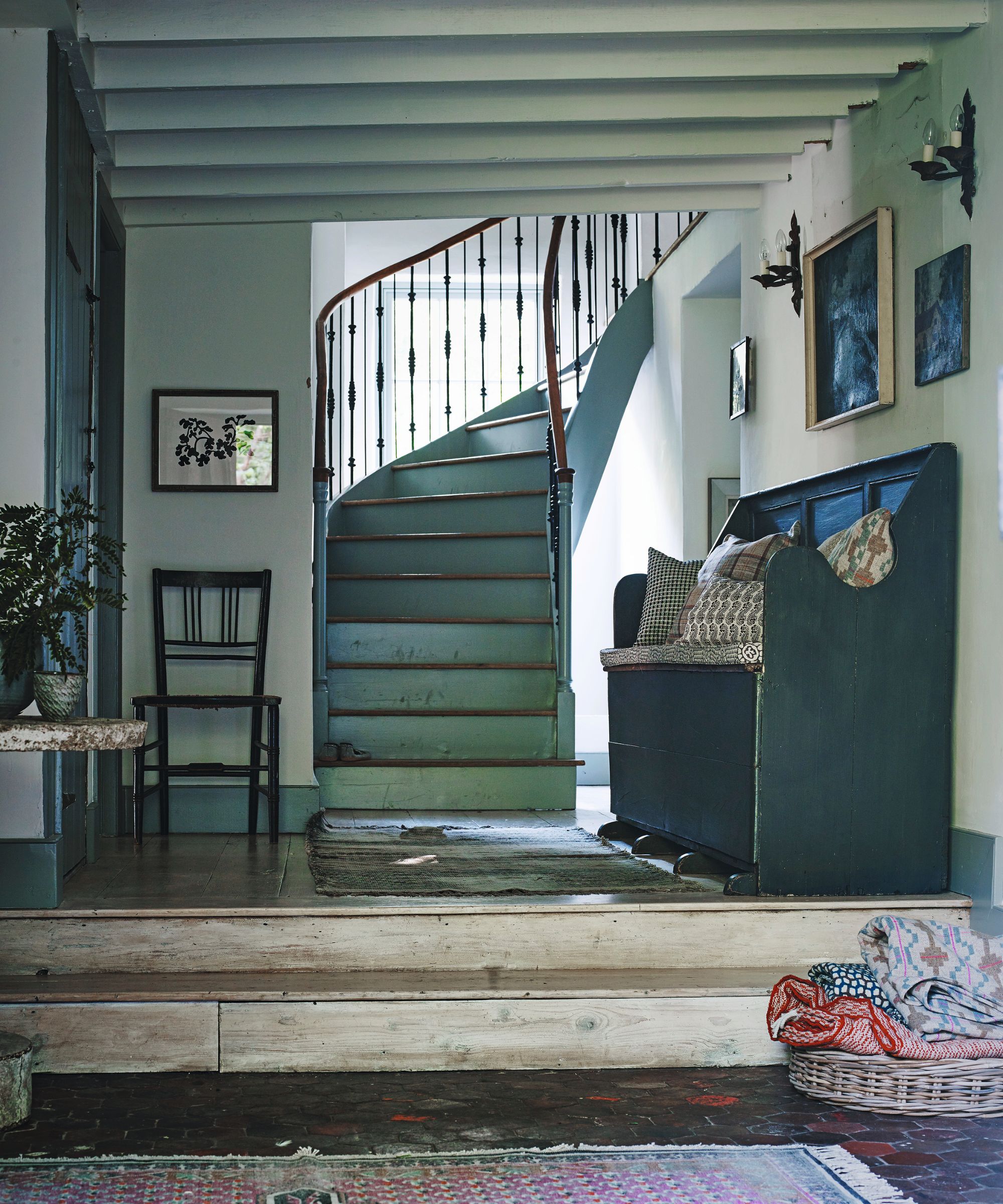 A blue, farmhouse style entryway with light worn wooden steps and panelled floor up to a curved blue staircase with a wooden banister. To the right is a wooden bench seat with patterned cushions below framed artwork, and to the left is a wooden chair.
