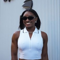 a black woman wearing a sleeveless white shirt and black sunglasses smiling in front of a white background