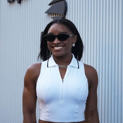 a black woman wearing a sleeveless white shirt and black sunglasses smiling in front of a white background