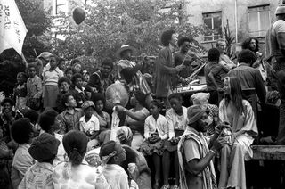 Black and white photograph of a large group of children sat on a float, some holding balloons while a number of adults are stood by, with some playing musical instruments.