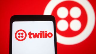 A phone shows the Twilio logo in red, while in the background a large Twilio logo in white is displayed on a wall